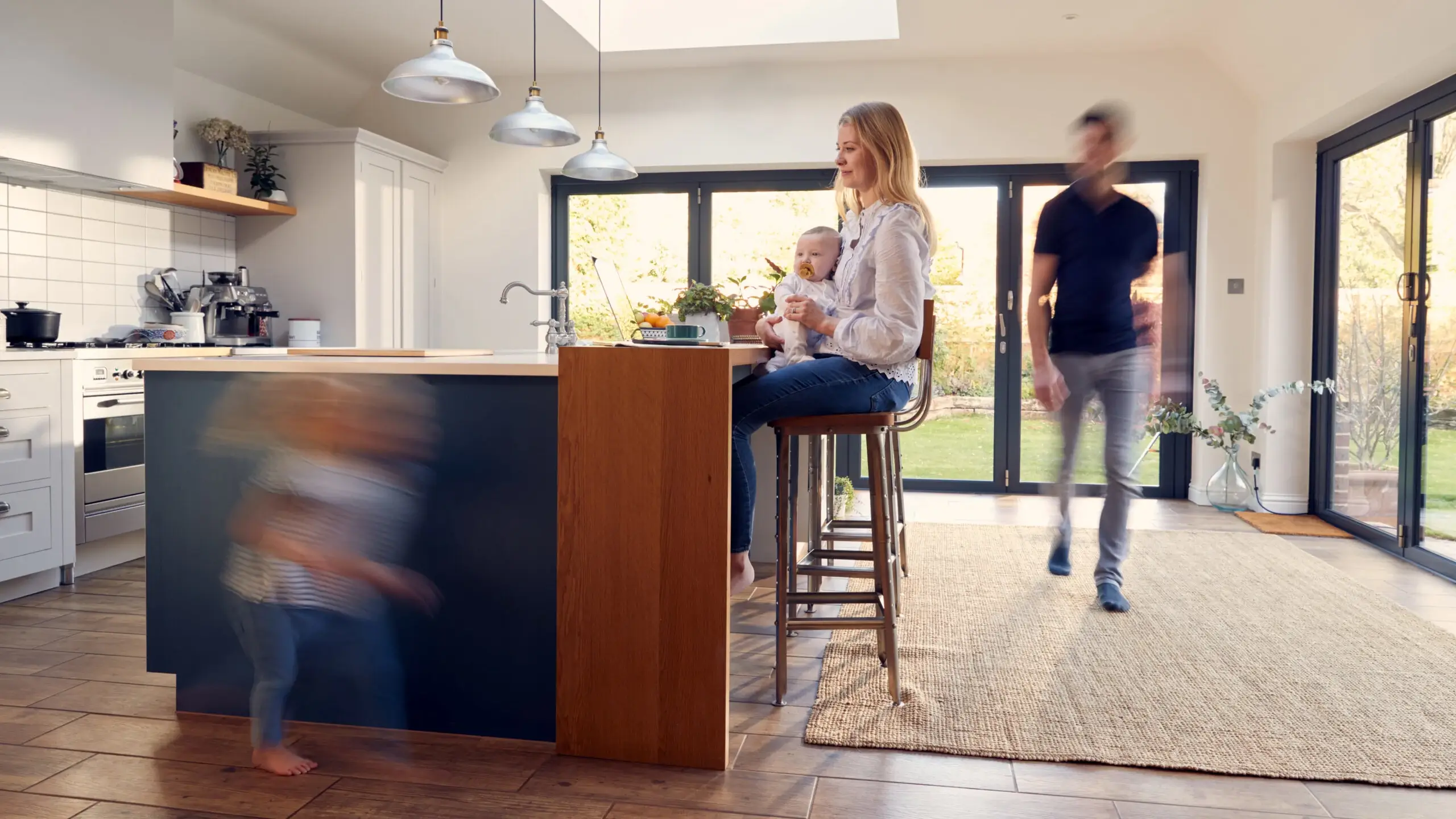 Woman sits at kitchen island with a baby, blurred man walks by, and child runs in the foreground.